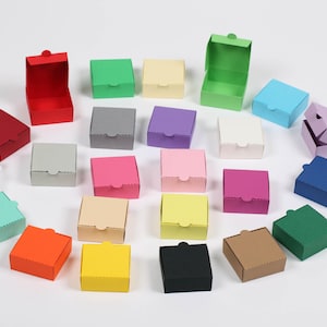 Könnte beinhalten: Eine Sammlung kleiner, quadratischer Geschenkboxen in verschiedenen Farben, darunter Rot, Grün, Blau, Gelb und Rosa. Einige Boxen sind geöffnet und zeigen ihr Inneres. Die Boxen sind auf einer weißen Oberfläche angeordnet.