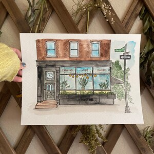 May include: Watercolor painting of a storefront with a black awning and a sign that says "79 Kenny St." The storefront has large windows with the words "Breakfast", "Lunch", and "Dinner" written on them. There are yellow lights hanging from the ceiling inside the storefront. The building is brick with three windows on the top floor.