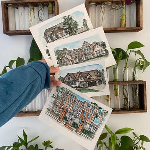 May include: Four watercolor paintings of houses, each framed and displayed on a wall with potted plants. The paintings are in a light blue, brown, and green color scheme.