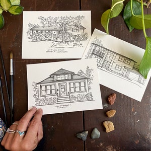 May include: Three black and white ink drawings of houses on white card stock. The drawings are on a wooden table with a vase of flowers and some small rocks.