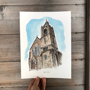 May include: Watercolor painting of a church with a tall tower and a bell. The church is painted in shades of brown and gray, with a blue sky in the background. The painting is signed "April Es Sold" in the bottom right corner.