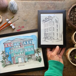 May include: Two framed watercolor paintings of houses. The painting on the left is a colorful depiction of a beach house with a red door and a palm tree. The painting on the right is a black and white line drawing of a city house with multiple windows.