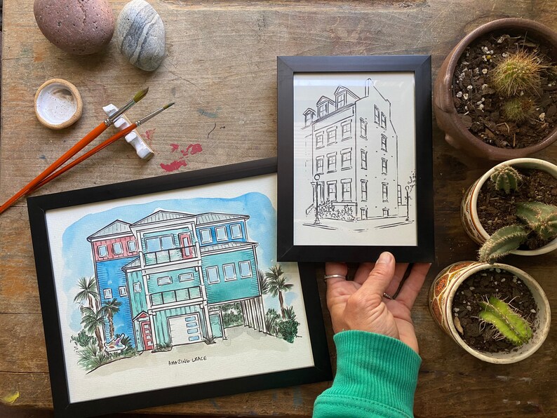 May include: Two framed watercolor paintings of houses. The painting on the left is a colorful depiction of a beach house with a red door and a palm tree. The painting on the right is a black and white line drawing of a city house with multiple windows.