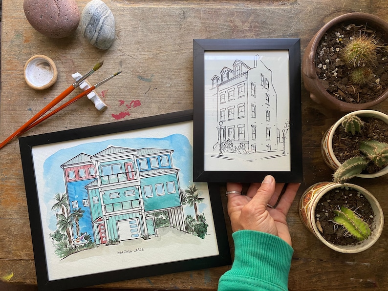 May include: Two framed watercolor paintings of houses. The painting on the left is a colorful depiction of a beach house with a red door and blue and green siding. The painting on the right is a black and white line drawing of a city house with a white door and black windows.