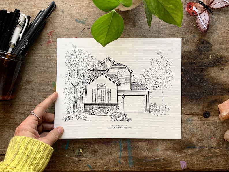 K&ouml;nnte beinhalten: Schwarz-wei&szlig;-Zeichnung eines Hauses mit Garage, B&auml;umen und einer Stra&szlig;enlaterne. Das Haus hat eine Backsteinfassade und ein Satteldach. Die Zeichnung ist auf wei&szlig;em Hintergrund.