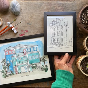 K&ouml;nnte beinhalten: Zwei gerahmte Aquarellbilder von H&auml;usern. Das Bild links ist eine farbenfrohe Darstellung eines Strandhauses mit einer roten T&uuml;r und blauer und gr&uuml;ner Verkleidung. Das Bild rechts ist eine Schwarzwei&szlig;-Zeichnung eines Stadthauses mit einer wei&szlig;en T&uuml;r und schwarzen Fenstern.