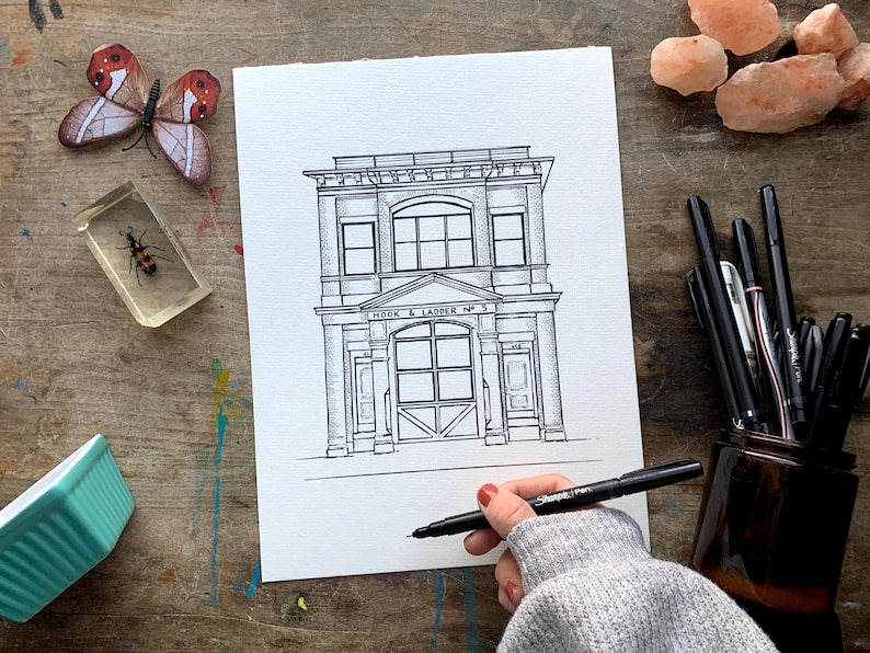 May include: Black and white line drawing of a building with the text "Hook & Ladder No. 5" above the entrance. The building has a large garage door and two windows on each side.