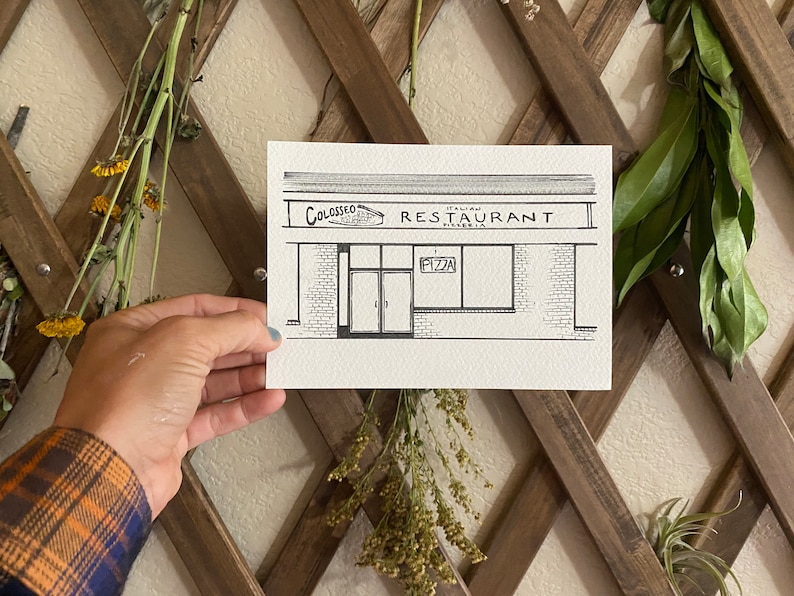 May include: A black and white line drawing of a restaurant storefront with the name "Colosseo Italian Restaurant Pizzeria" above the entrance. The storefront has a brick facade and two large windows. A sign above one of the windows reads "Pizza."