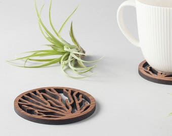 May include: Two wooden coasters with a tree branch design. The coasters are brown and have a natural wood finish. A white ceramic mug is sitting on one of the coasters. Two small green air plants are on the table.