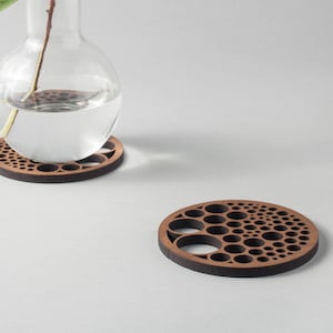 Puede incluir: Dos posavasos de madera con un diseño circular que presenta un patrón de círculos de varios tamaños. Un jarrón de vidrio transparente con un solo tallo de hojas verdes se encuentra sobre uno de los posavasos.