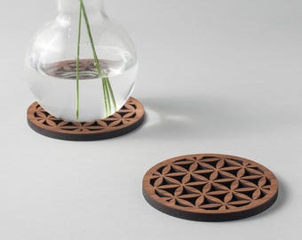 May include: Two wooden coasters with a flower of life design. The coasters are brown and have a intricate pattern. A clear glass vase with lavender flowers sits on one of the coasters.
