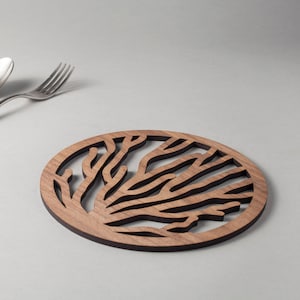 以下が含まれることがあります： 枝分かれしたサンゴの模様がくり抜かれた丸い木製コースター。コースターは薄い茶色の木でできており、濃い茶色の縁取りがあります。