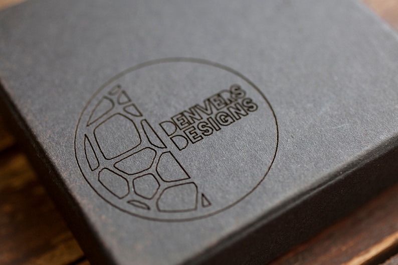 May include: A close-up of a black box with a circular logo. The logo features a geometric design and the text "DENVERS DESIGNS".