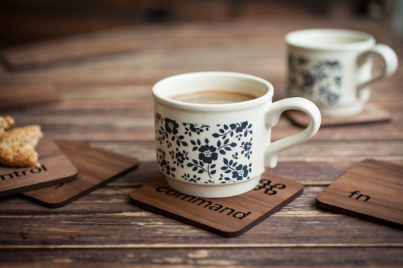 May include: Two white ceramic mugs with black floral patterns, one filled with coffee, sitting on wooden coasters with the words "control", "command", "fn" engraved on them.