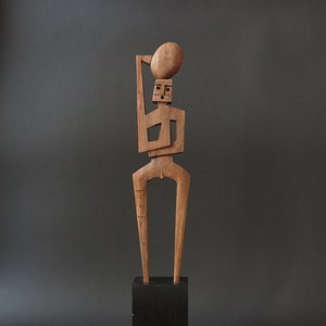 Könnte beinhalten: Eine Holzskulptur einer stilisierten menschlichen Figur, die ein rundes Objekt über ihrem Kopf hält. Die Figur steht auf einem schwarzen Holzsockel.