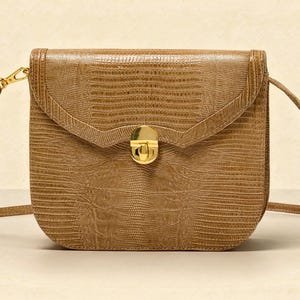 Könnte beinhalten: Braune, strukturierte Umhängetasche mit Klappenverschluss und goldfarbener Hardware. Die Tasche hat einen langen, verstellbaren Riemen und eine rechteckige Form. Das Design der Tasche umfasst eine dekorative Klappe und einen goldfarbenen Verschluss.