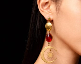 May include: Gold-toned drop earrings with a celestial theme. Each earring features a small ball stud, a circular disc, a red oval gemstone, and a crescent moon with a sunburst design. The earrings are worn on a person's ear.