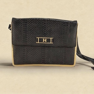 Può includere: Borsa a tracolla nera con un motivo strutturato a forma di serpente. La borsa presenta una cornice color oro e un accento rettangolare dorato con la lettera "H". È attaccata una lunga tracolla nera.
