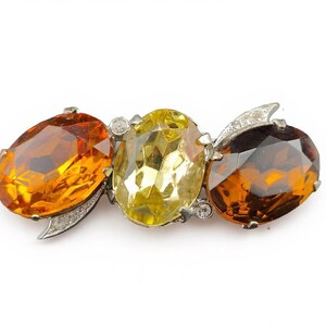 May include: A vintage brooch with three large, oval gemstones in amber and yellow hues. The stones are set in silver-toned metal with small accent stones and decorative flourishes.