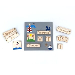 May include: A wooden calendar with a gray background and colorful illustrations. The calendar includes months, days of the week, and numbers. The calendar is designed for children and features a snowman, a lighthouse, a sun, and an anchor.