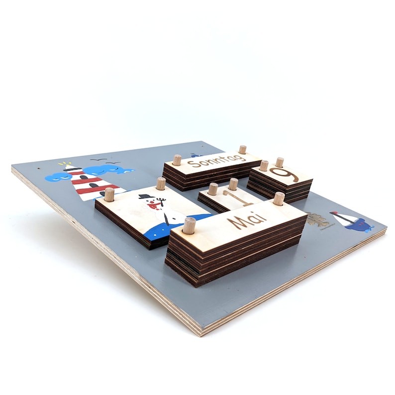 May include: Wooden calendar with a gray background and a lighthouse, snowman, and sailboat design. The calendar features a date display with the numbers 1 and 9, the month "Mai" (German for May), and the day of the week "Sonntag" (German for Sunday).