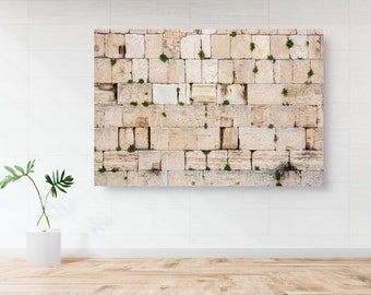 May include: A close-up of a large stone wall with small green plants growing in the cracks. The wall is made of light brown and beige stones, and the mortar is a lighter shade of beige. The wall is very old and weathered, and the stones are uneven in size and shape.
