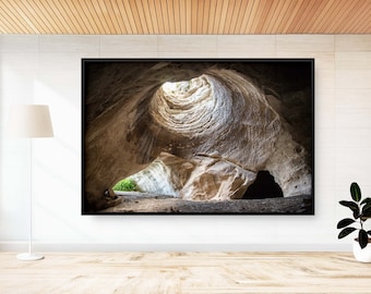 May include: A black framed photograph of a cave opening with a circular opening at the top, revealing a bright light. The cave walls are a light brown color and the opening is surrounded by a dark brown rock formation.