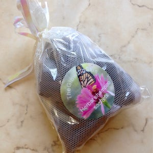 May include: A white mesh bag with a pink and white ribbon tied around the top. The bag contains several brown herbal shampoo sachets. The bag has a round label with a butterfly and flowers, and the text "The Glow Skin Herbal shampoo sachets".