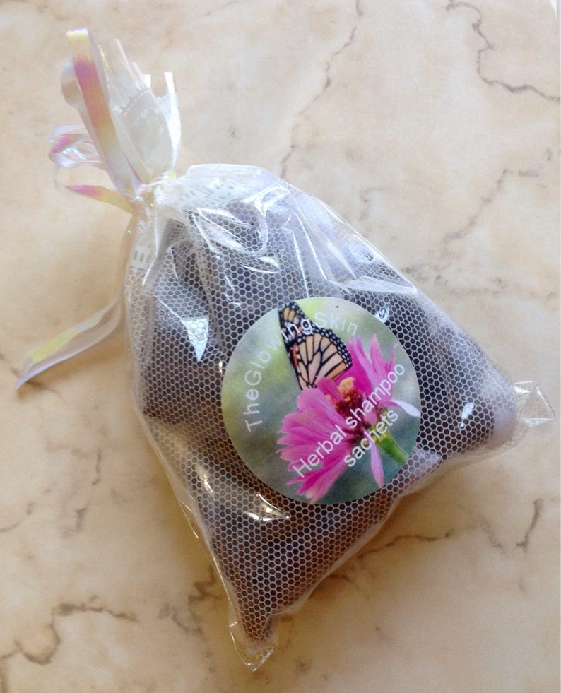 May include: A white mesh bag with a pink and white ribbon tied around the top. The bag contains several brown herbal shampoo sachets. The bag has a round label with a butterfly and flowers, and the text "The Glow Skin Herbal shampoo sachets".