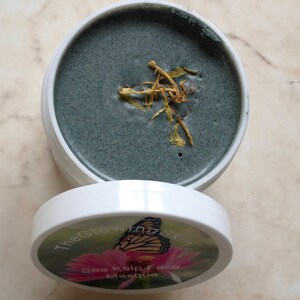 May include: A white jar with a black lid. The lid has a label that says "The Glowing Skin Sea Kelp Face Masque" and features a butterfly on a pink flower. The jar is open and contains a dark gray face mask with dried yellow flower petals on top.