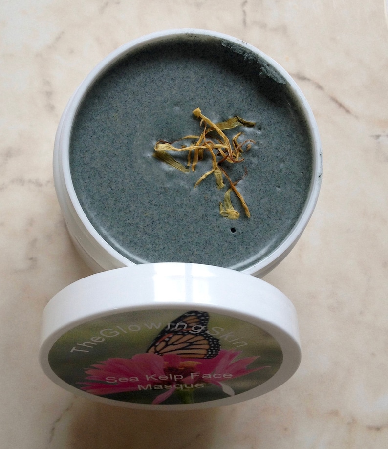 May include: A white jar with a black lid. The lid has a label that says "The Glowing Skin Sea Kelp Face Masque" and features a butterfly on a pink flower. The jar is open and contains a dark gray face mask with dried yellow flower petals on top.