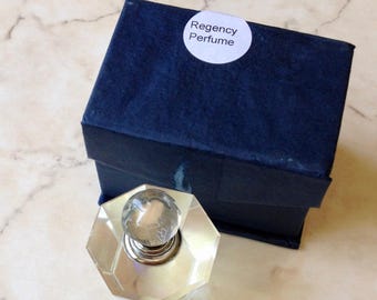May include: A clear glass perfume bottle with an octagonal base and a spherical stopper, positioned in front of a dark blue rectangular box. A white circular sticker on the box lid features the black printed text Regency Perfume.
