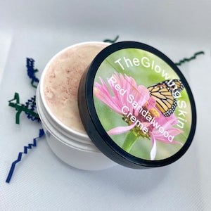 May include: A white jar with a black lid containing a red sandalwood creme. The lid has a green and pink flower with a butterfly on it. The text on the lid reads "The Glow in the Skin Red Sandalwood Creme".