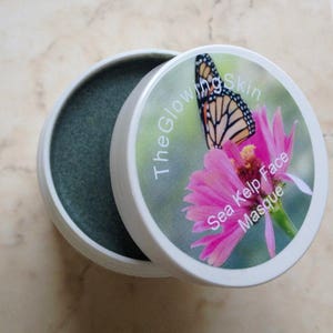 May include: A white jar with a green face mask inside. The lid of the jar has a pink flower with a butterfly on it. The text on the lid reads "The Glowing Skin Sea Kelp Face Masque."