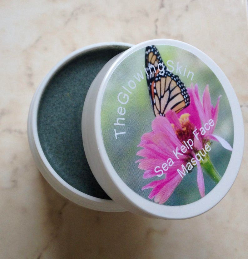 May include: A white jar with a green face mask inside. The lid of the jar has a pink flower with a butterfly on it. The text on the lid reads "The Glowing Skin Sea Kelp Face Masque."