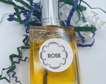 May include: A small, square glass bottle filled with a yellow liquid, topped with a silver cap. A white label with the word "ROSE" is affixed to the bottle. The bottle is surrounded by green, blue, and white paper shreds.