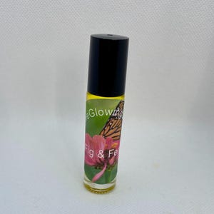 Puede incluir: Un frasco roll-on de vidrio transparente con tapa negra. La etiqueta presenta una flor rosa, una mariposa monarca y las palabras "eGlowing" y "Fig & Fel". El frasco contiene un líquido amarillo.