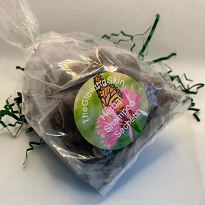 May include: A clear plastic bag filled with brown herbal shampoo sachets. The bag has a round sticker with a butterfly, flowers, and the text "The Glowing Skin Herbal Shampoo Sachets".