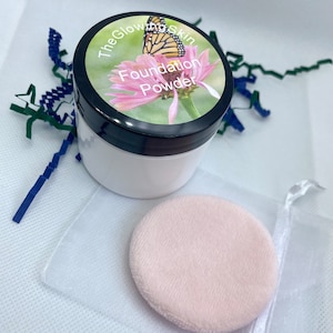 Może przedstawiać: Biały słoik z czarną pokrywką i etykietą z napisem "The Glowing Skin Foundation Powder". Etykieta przedstawia motyla i różowy kwiat. W zestawie znajduje się różowy puder w białej siateczkowej torebce.