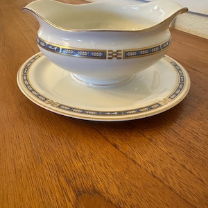 May include: A white porcelain gravy boat and matching saucer. The gravy boat has a curved spout and a handle. Both pieces are decorated with blue and gold bands and geometric patterns. The set is placed on a wooden surface.