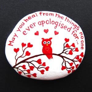May include: A white painted stone featuring a red owl perched on a brown branch with red heart-shaped leaves. The text "May you heal from the things no one ever apologised for..." is written in red lettering above the owl with three small red hearts.