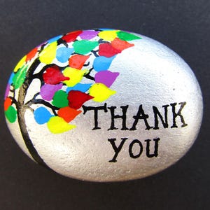 May include: A smooth, silver-painted river stone featuring a hand-painted black tree silhouette with vibrant, multi-coloured leaves in red, yellow, blue, green, and purple. The words THANK YOU are printed in black capital letters on the right side.