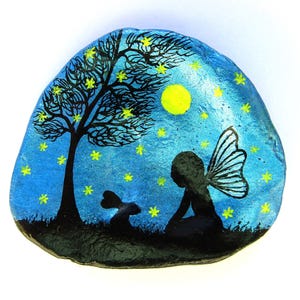 May include: A hand-painted rock depicting a night scene. A black silhouette of a tree, fairy, and rabbit are set against a blue background with yellow stars and a yellow moon. The rock is an irregular shape.