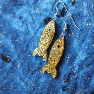 May include: A pair of gold glitter rocket ship earrings with silver hooks. The earrings are on a blue fabric background.