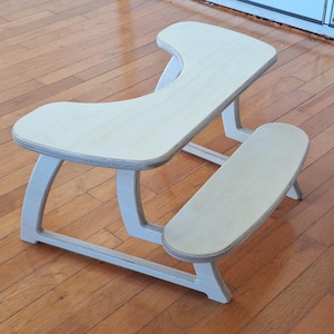 May include: A light-colored wooden kneeling chair with a curved seat and a separate knee rest. The chair has a minimalist design with white supports and is designed for ergonomic posture.