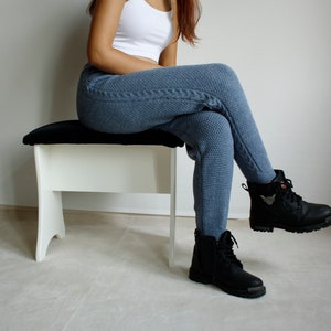 Woman seated on a white bench wearing denim blue cable leggings, showing the textured knit pattern from hip to ankle.