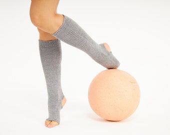 May include: A person wearing gray leg warmers with a hole at the toe balances on a large, round, peach-colored ball.