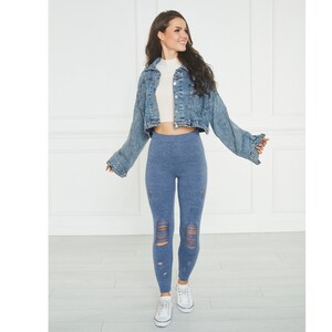 Puede incluir: Una chaqueta corta de mezclilla azul con mangas largas, una blusa blanca de cuello alto, leggings azules desgastados con roturas horizontales en las rodillas y espinillas, y zapatillas blancas con cordones. El conjunto tiene un estilo casual.