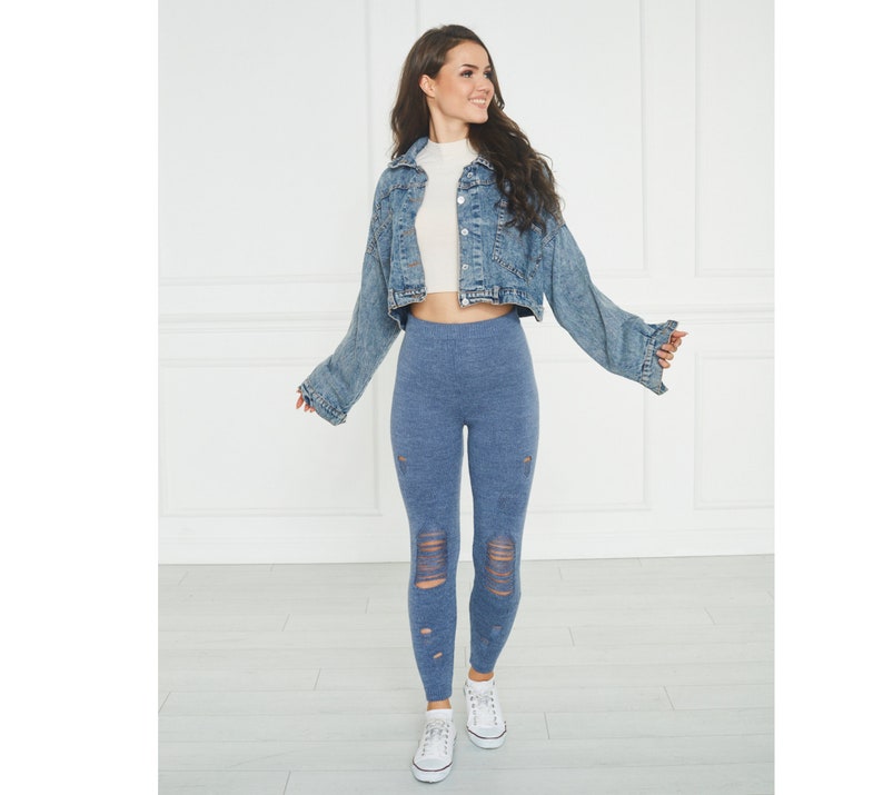 Puede incluir: Una chaqueta corta de mezclilla azul con mangas largas, una blusa blanca de cuello alto, leggings azules desgastados con roturas horizontales en las rodillas y espinillas, y zapatillas blancas con cordones. El conjunto tiene un estilo casual.