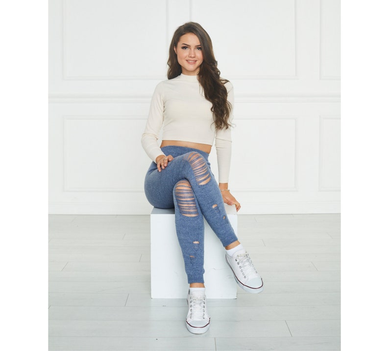 Puede incluir: Un top corto de color crema con mangas largas y cuello alto combinado con leggings azules desgastados que presentan rasgaduras horizontales a lo largo de las piernas y zapatillas blancas con cordones. Los leggings tienen un aspecto tejido.